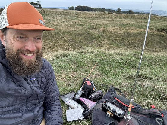Selfie of guy with beard and orange cap smiling, sitting on the grass with a pile of radio gear. KX2, quansheng.