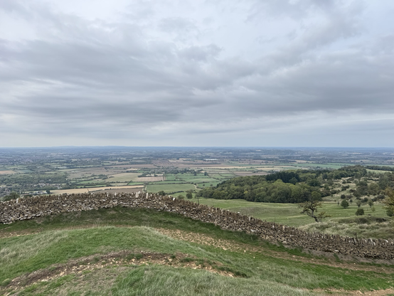 Hill top view with a patchwork of flat field down below in the distance and a dry stone wall that undulates across the foreground.