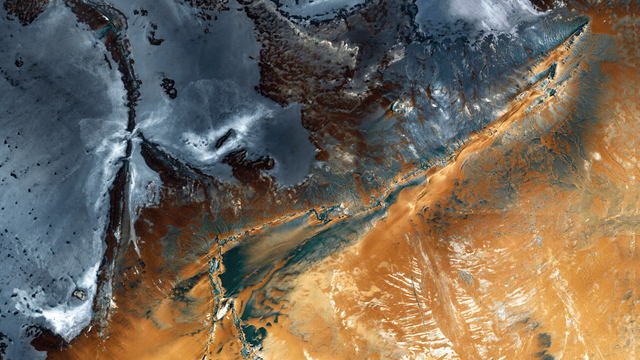 A satellite picture of a mountainous desert landscape. The upper and central areas feature rugged mountain ridges, while the lower right displays sandy dunes and eroded valleys in warm brown and orange tones.