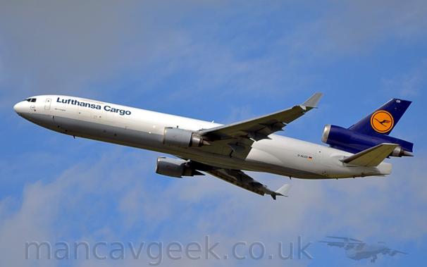 Side view of a 3 engined jet airliner flying from right to left at a low altitude, with undercarriage already tucked away, flaps extended from the rear of the wings, and the nose raised significantly, suggesting that it has just taken off.
The plane is mostly white, with a grey belly, and dark blue "Lufthansa Cargo" titles on the upper forward fuselage, and the black registration "D-ALCD" next to a small German flag on the lower rear.
The tail and centre engine pod are dark blue, with a yellow circle containing a stylised blue flying bird in the middle.
The engine pods mounted under the wings are grey, as are the up-turned wing-tips.
Pale blue sky with grey fluffs of cloud fill the rest of the frame.