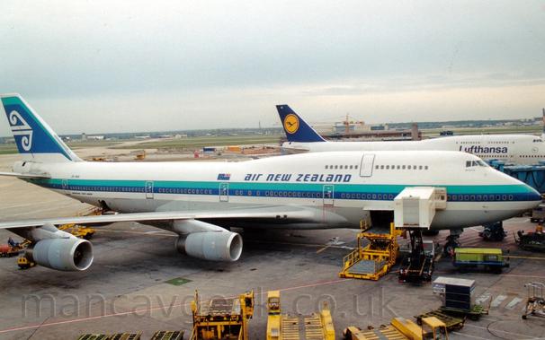 Side view of a very large, 4 engined jet airliner parked at an airport terminal, facing to the right.
The plane is mostly white, with an aquamarine and blue stripe running along the body, the blue part covering the passenger windows on the main deck.
There are blue "Air new Zealand" titles on the upper forward fuselage, next to a New Zealand flag.
The tail is mostly blue, outlined with a thin aquamarine stripe on the top and white at the bottom, with a pair of stylised white waves in the middle.
Yellow baggage loaders litter the grey concrete ramp in the foreground, some connected to open hatches in the lower fuselage.
In the background, another very large jet airliner with a similar shape, but with a dark blue tail with a dark blue stylised flying bird in a yellow circle in the middle, with dark blue "Lufthansa" titles on the upper forward fuselage.
Beyond that plane, a general view across the airfield sees extensive areas of green grass separated by taxiways and runways, with planes and buildings in the distance, under grey skies.