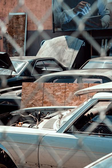 Cars in various states of disrepair are crammed in a cluttered lot. The hand of an executive pictured in a billboard looms above.