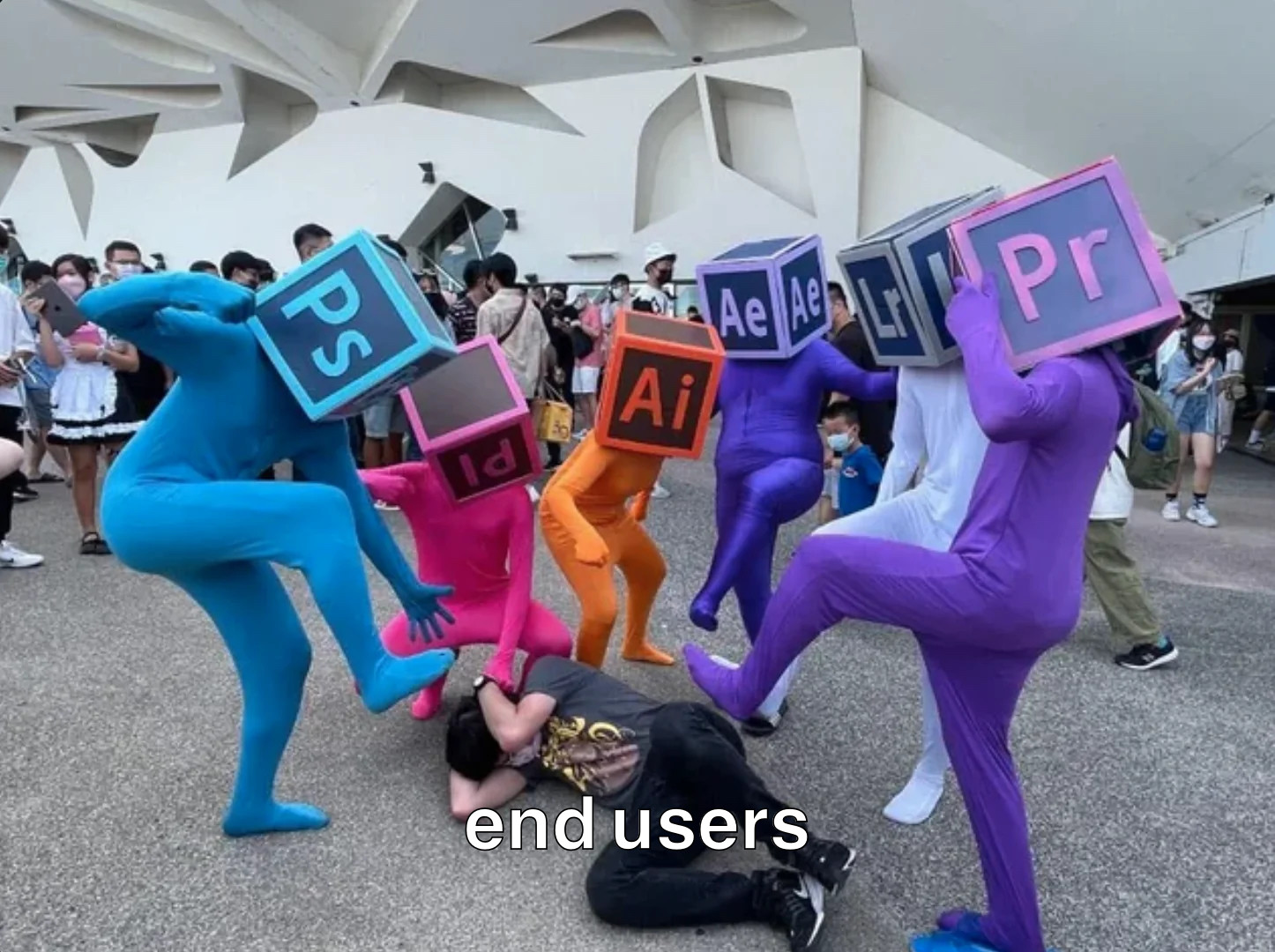 A group in multicoloured Adobe Suite themed lycra suits (wearing square head mask for the icons for: Photoshop, Prelude, Illustrator, After Effects, Lightroom, & Premiere Pro.) collectively beating on a person bracing on the ground (Captioned End User). Original uncaptioned photo via: u/Cybernaut