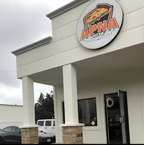 A beige storefront with pillars. Above the shop is sign "Apna Pizza" with a cartoon slice of pizza