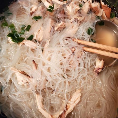 Close-up overhead view of a bowl of chicken noodle soup. The soup features thin, translucent noodles, shredded chicken, and chopped green onions in a clear broth. Wooden chopsticks rest within the soup bowl.