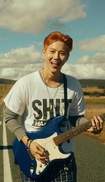 Orange haired kpop star SUHO plays his blue electric guitar on a highway somewhere in Australia, wearing a white short sleeved shirt that says "SHIT HAPPENS" over a gray long sleeved shirt. He is grinning while sticking his tongue out.