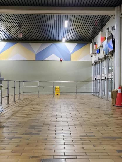 Showing an empty ferry terminal, with brown 1970s tiling, a metal railing leading to the doors to the pier. There is a beige/green wall, with a wet floor sign leaning against it, and some blue and yellow decorations above the wall with some lights shining down