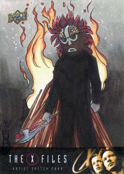 Image is of a traditional illustration done on a licensed trading card blank. The bottom of the card is decorated with the set logo: The X-Files, the text Artist Sketch Card and black and yellow graphic under the logo and text. The illustration is a scene from an X-Files episode. In the foreground is a figure wearing a tribal styled wooden mask that has a fringe of blood red feathers. He holds an axe in his right hand, and stands in front of a roaring bonfire. The art is signed DG 25 in the lower corner, the initials for the artist Diana Greenhalgh and the year the art was produced for the set. The art is rendered with Copic markers with deep shadows in the foreground and soft shadows in the background.