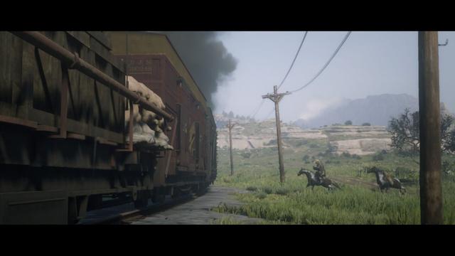 The screenshot depicts a western-themed game environment. In the foreground, there is a dirt path that leads to a train station with two old trains on rails. One of the trains appears to be an engine pulling freight cars, while the other seems to be an empty passenger car. There are no visible people or animals on the ground near the trains. The background features a rural landscape with rolling hills and sparse vegetation under a hazy sky. The scene suggests an older time period in a western setting, possibly indicating that it is part of the storyline set within Red Dead Redemption 2.