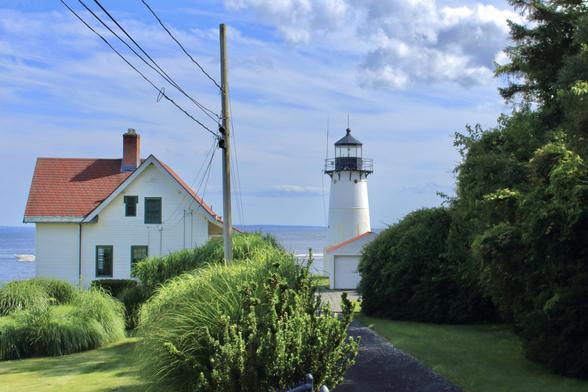 A white lighthouse with a black lantern stands near the shoreline, next to a white house with a red roof. A narrow paved path leads through green bushes and grass toward the lighthouse and the ocean in the background. Power lines cross the view overhead.