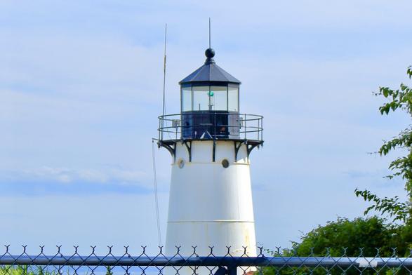 White lighthouse with black lantern room and green light, seen above a chain-link fence. The tower has two antennas and is surrounded by trees on the right and water in the background under a partly cloudy sky.