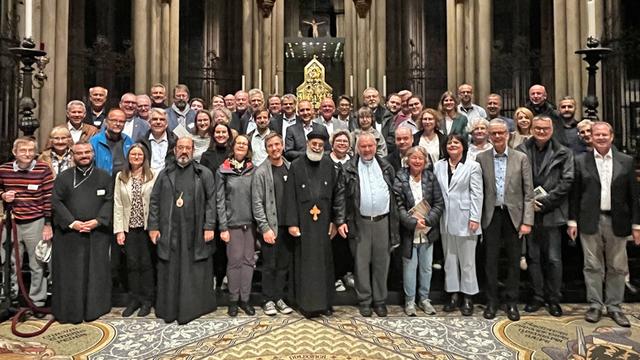 Das Gruppenfoto zeigt mehr als 50 Personen, die in einer prächtigen, hochgewölbten Kirche posieren. Die Gruppe besteht aus Männern und Frauen unterschiedlichen Alters, viele davon in formeller oder semi-formeller Kleidung. Einige Personen in der vorderen Reihe tragen religiöse Gewänder, darunter ein Mann mit einem langen schwarzen Gewand und einem weißen Bart, der in der Mitte steht. Der Hintergrund ist eine beeindruckende gotische Kirchenarchitektur mit hohen Säulen, Spitzbögen und Statuen in Nischen.