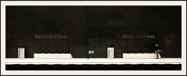 Das Bild zeigt eine U-Bahn-Station in Berlin – „Rotes Rathaus“ als Stationsname ist gut sichtbar. Die Wände sind aus dunklem, glänzendem Material, der Boden hell. In der linken und rechten Bildhälfte sind je ein Mülleimer, eine Bank und der Stationsname an der Wand sind zu sehen. In der Mitte ist ein grünes Fluchtweg-Schild mit einem nach rechts zeigenden Pfeil angebracht. Auf der Sitzbank in der rechten Bildhälfte sitzt eine Person auf dem Platz ganz rechts außen und damit am rechten Bildrand. Sie hat die Beine verschränkt und scheint auf ihr Handy zu schauen. Der allgemeine Eindruck ist ruhig und minimalistisch. Das Bild wirkt kühl und funktional, beinahe steril.

The picture shows a subway station in Berlin – “Rotes Rathaus” (Red Town Hall) is clearly visible as the station name. The walls are made of dark, shiny material, and the floor is light-colored. On both the left and right sides of the picture, there is a trash can, a bench, and the station name on the wall. In the middle, there is a green emergency exit sign with an arrow pointing to the right. On the bench on the right-hand side of the picture, a person is sitting on the seat on the far right, at the edge of the picture. They have their legs crossed and appear to be looking at their cell phone. The overall impression is calm and minimalistic. The picture appears cool and functional, almost sterile.