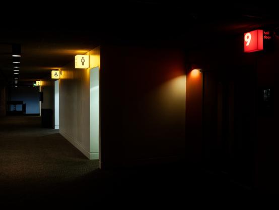 Dark corridor with square lights designating toilets and a room. It's all very dark and empty.