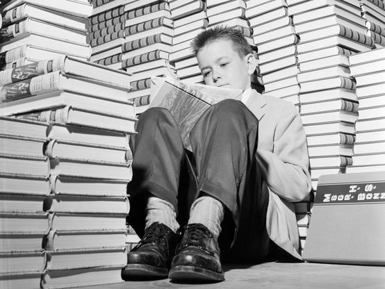 Un niño lee en una biblioteca junto a una pila de libros en los años cincuenta.