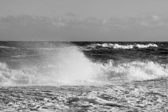 A black and white photo of a large wave crashing violently onto a beach, creating a large plume of spray and foam. The water is a dark shade and the sky above is cloudy, giving the image a dramatic and moody feel. Smaller waves are visible in the background, rolling in from the dark horizon.