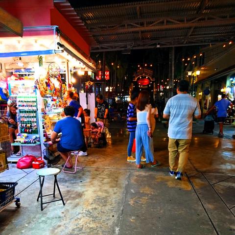 Eine Straßenszene in Bangkok, Thailand, bei Nacht. Im Vordergrund die durch den Laden links beleuchtete Straße mit mehreren Bummelanten. Im Hintergrund dunkle Nacht. Oben ein Metalldach.
