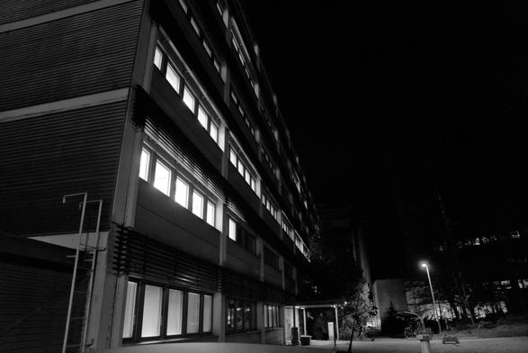 A black and white photograph of the side of an empty office building. Light come out from inside, shining though every window, arranged as a regularly spaced row of rectangles at every floor. The wide angle of the lens give the building an imposing presence.