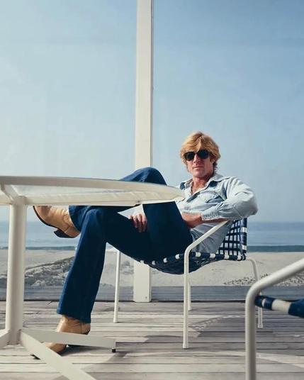 Redford in a jeans shirt and jeans trousers with light brown boots. Sitting in a loungey chair. Sunglasses, beach in the back.