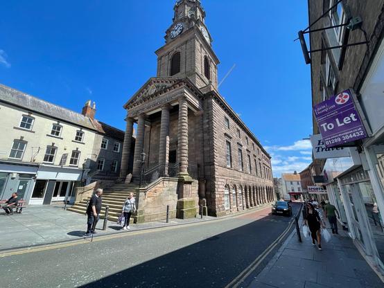 Under a vivid blue sky, the historic Town Hall dominates Marygate in Berwick-upon-Tweed, its grand stone façade and clock tower rising above the street. The neoclassical columns and wide steps lend an imposing presence, while intricate details along the pediment reflect the town’s rich heritage. Beside it, pale terraces with pitched roofs and chimneys line the square, some with shuttered windows and quiet shopfronts, evoking a sense of timeless charm. On the opposite side, modern shop signs, including a “To Let” notice, hint at the blend of old and new life along the thoroughfare. Pedestrians stroll along the pavement, some carrying shopping bags, while a few pause near the bollards and benches that edge the street. A row of arched windows along the side of the hall stretches toward the distance, drawing the eye down the gently curving road where sunlight illuminates the red brick roofs further along. The street feels calm and inviting, with light and shadow playing across the stonework and pavement, creating a warm, late-morning atmosphere in the heart of the town.