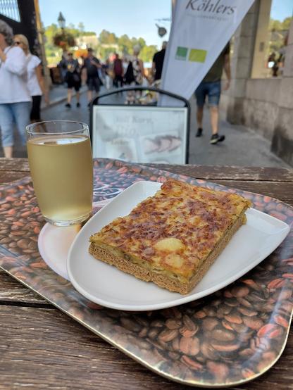 Auf einem Stehtisch aus Holz liegt auf einem Teller ein Stück vegetarischer Zwiebelkuchen. Daneben steht ein Glas mit Federweißer. Im Hintergrund erkennt man die Alte Mainbrücke in Würzburg.