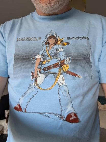 A light blue t-shirt with a printed image of Princess Nausicaa in the centre standing with her legs slightly apart, holding a large rifle and with a determined look on her face. Tope left is the lettering NAUSICAÄ and top right is the Japanese lettering 風の谷のナウシカ.