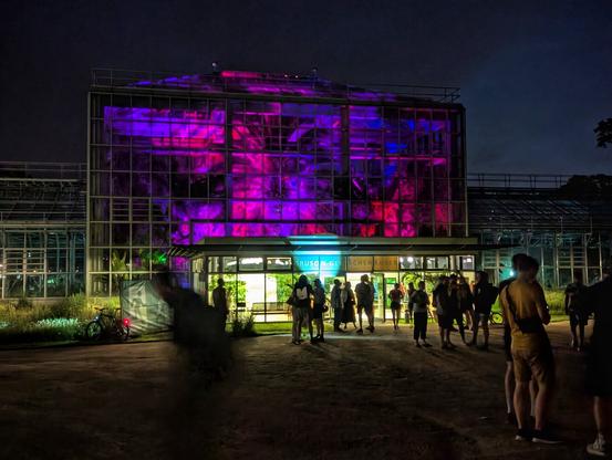 A vibrant night-time shot of a large glass building, possibly a botanical garden or conservatory, illuminated with striking pink and purple lights. A crowd of people are gathered in front of the entrance, their figures silhouetted against the bright interior. The building features multiple levels and numerous window panes, reflecting the colourful light. The ground in front appears to be a paved area, with some shadowy figures and a parked bicycle visible on the left. The sky above is dark, adding to the dramatic contrast of the illuminated structure.