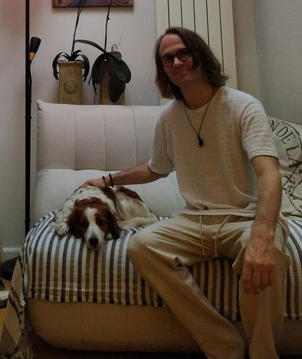 A photo in a light white setting with a dog lying relaxed on the left-side of a white couch and a guy with long hair and glasses (me) sitting on the right, looking into the camera, and petting the dog.