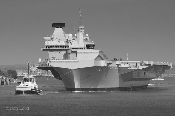 This is a black and white maritime photo of an aircraft carrier leaving harbour and undertow by a immaculately clean, large tug.
In the foreground and rising upwards about one third of the image is a mass of dark grey and calm water. In the lower left corner is a large tug underway from right to left. The tug is clean, smart with a black hull but with a large white wrap-around fender on the bow with two parallel white rubbing strakes surrounding the hull. On top is a 'control tower' like wheelhouse with large darkened windows and a tall signal mast on top with numerous antennae. A short distance behind the tug, about ten metres and attached with a taut and stout line is an aircraft carrier. The vessel is rendered a light grey and lit by the bright sun light. The flight deck overhangs the hull on both side. On the port side and over the bow is the distinctive ski-jump ramp. On the starboard side of the flight deck are the two 'islands', the forward one for ship operations the aft one for aerial operations. On either side of the flight deck are a few of the crew. Behind the carrier is the scene of a large harbour with many moored sailing boats. Above is a clear and cloudless sky rendered a medium grey.
The ship: HMS Queen Elizabeth and the location: Portsmouth, UK (2019).