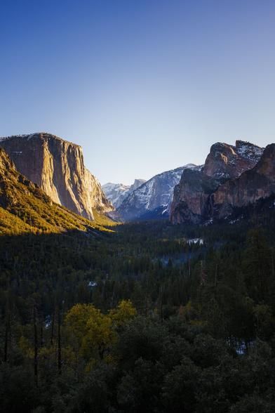 A panoramic view of a valley surrounded by towering mountains, bathed in warm sunlight under a clear blue sky.