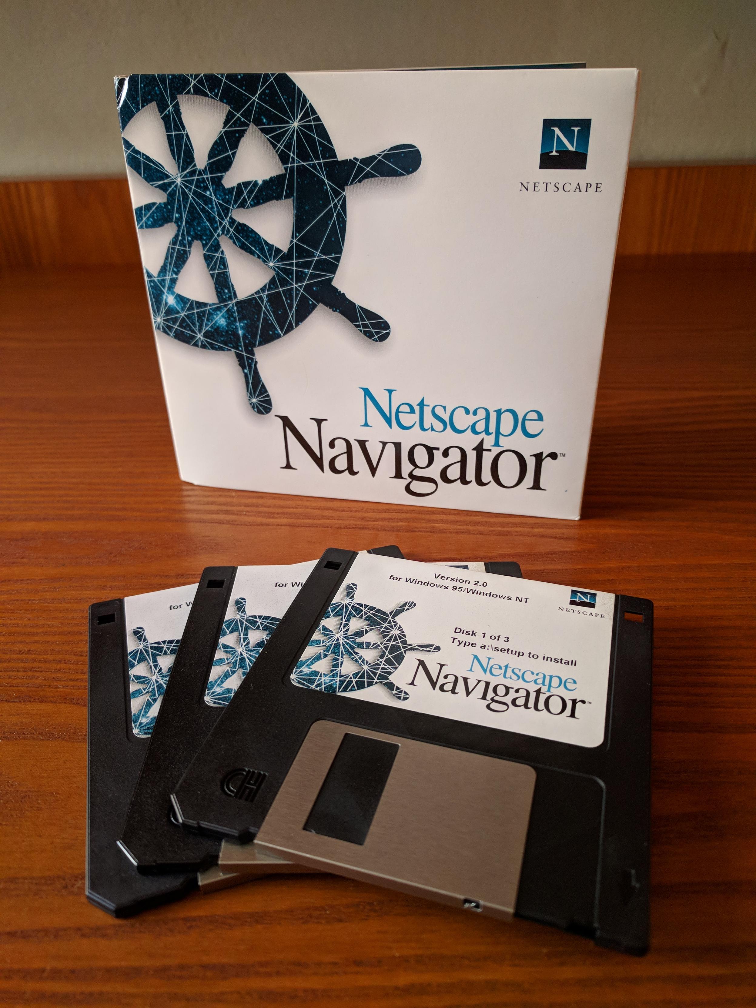 A photo of 3 black 3.5" micro floppy disks spread as a fan on a wooden desk. Behind the disks, set vertically on the desk as an open book, is a cardboard sleeve with 3 slots for the disks. They are the distribution disks and sleeve of the Netscape Navigator 2.0 web browser for Windows 95 and NT. The disks have labels with white background and the sleeve has a cover with a white background. Along with the product name and version, the labels and cover have the product logo and the artwork of a marine rudder wheel.
