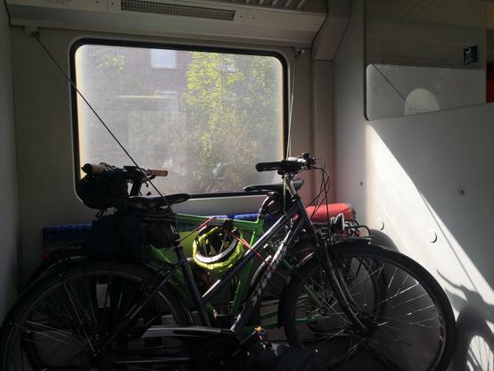 Zwei Fahrräder mit Gepäck stehen im Zug in einem Abschnitt mit Fenster, der kürzer als eine Fahrradlänge ist, nebeneinander und sind mangels Befestigungsmöglichkeiten mit vier Spanngurten an der Gepäckablage darüber befestigt. An der Trennwand rechts im Bild ist ein blaues Hinweisschild mit Kinderwagen und Fahrrad darauf zu sehen.