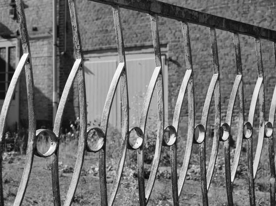 Rusty metal fence with a pattern of curved bars and circular cutouts, set in front of a brick building. A closed door and window are visible in the building's facade. Shadows are cast on the ground, indicating bright sunlight. The image is in black and white.