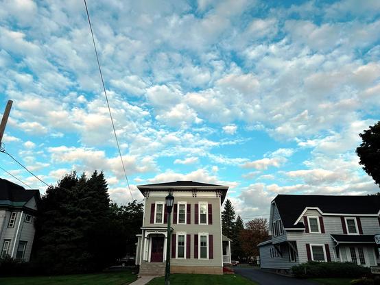 Fluffy white clouds congregate in a turquoise sky. Below are several houses, similarly colored with gray siding, dark roofs, and white-framed windows with black shutters. Behind the houses are trees, and in front a single lamppost.