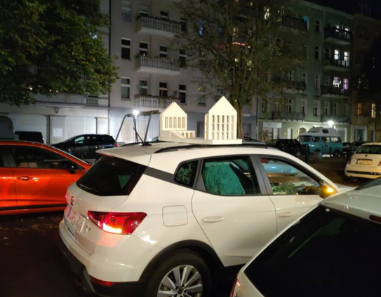 ein weißer Kleinwagen auf einer nächtlichen Straße. Auf dem Dach ist ein weißes Architekturmidell befestigt, sodass das Auto 2 Giebel zu haben scheint.