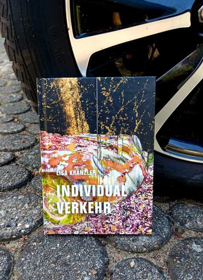 Foto des Buches, auf dessen Cover ein Ausschnitt aus einem Gemälde von Lisa Kränzler zu sehen ist: die Schnauze eines Autos in rundlichem Design, dessen Grundfarbe Weiß ist, das aber in anderen Farben bespritzt ist, als wäre es durch Matsch gefahren. Das Auto steht auf etwas, das an Herbstlaub erinnert, vor einem schwarzen Hintergrund, auf dem goldfarbene Strukturen Bäume und kahles Unterholz andeuten. Das Buch lehnt so an dem Rad eines Autos, dass nur dessen Reifen und ein Teil der Felge zu sehen ist.