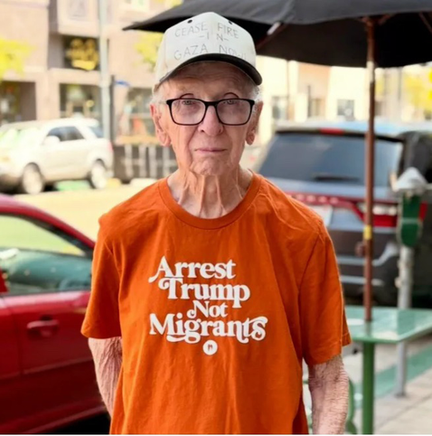 An elderly man wears a bright orange tee shirt outside near parked cars, that says Arrest Trump Not Migrants. 👍