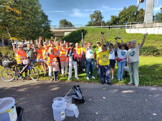 20 Menschen, die Ihre Müllsammelwerkzeuge in die Höhe recken, Warnwesten und T-Shirts anhaben. Wir lachen alle und freuen uns, trotz traurigem Anlass machen wir etwas sinnvolles zusammen an einem schönen Septembersamstag. Wir stehen auf einem Weg, hinter uns ist die Bundesstraße, die über eine Brücke geht, außerdem sieht man Grünfläche.