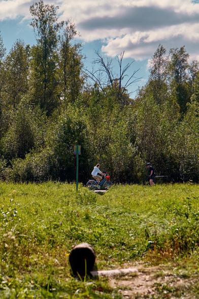 A cyclist rides through a grassy area, while a person walks nearby. Lush green trees and a cloudy sky serve as the backdrop. A colorful post is in the foreground.