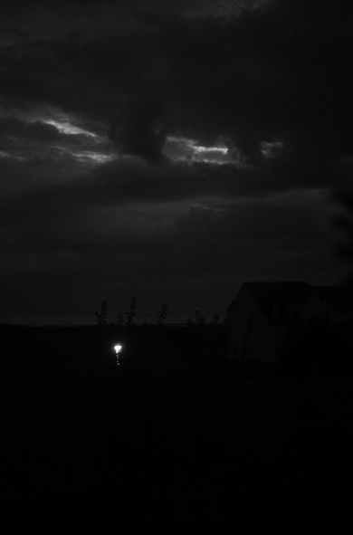 A black and white photography depicting a dark sky with a view on the sea. there is only one light visible