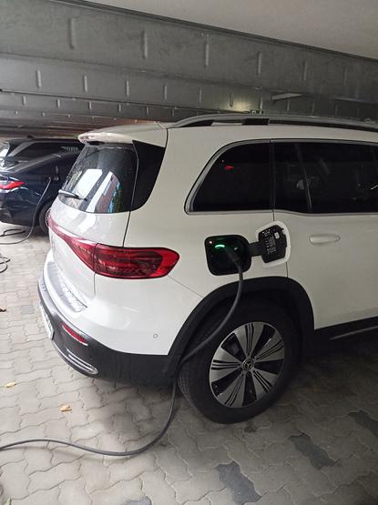 Unser Mercedes EQB 250+ in weiß beim Ladevorgang im Parkhaus des Wissenschaftsparks.
Man sieht das Heck rechts und das eingesteckte Kabel. 
Ladestatus leuchtet grün.