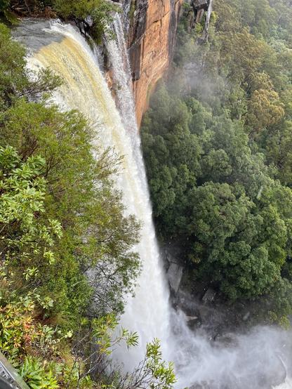 Fitzroy Falls, Australia Waterfall cascading over a sheer cliff, surrounded by dense green foliage. Mist rises from the base where the water crashes. The cliff has a reddish-brown hue, contrasting with the lush greenery.