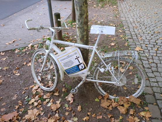 Ein weißes Ghostbike angelehnt an einem Baum nebem einem gepflasterten Gehweg. Zwei Schilder gedenken an Natenom / Andreas Mandalka.