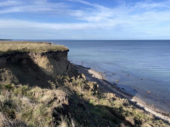 Eine Küstenszene mit einer grasbewachsenen Klippe mit Blick auf das ruhige Meer. Die Küstenlinie ist sichtbar, mit verstreuten Steinen entlang des Wasserrands unter einem klaren blauen Himmel mit einigen Wolken.
