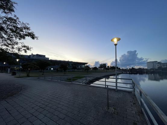 Eine ruhige Uferszene in der Abenddämmerung mit einem von Bäumen gesäumten Weg, einem modernen Gebäude und einem sanft beleuchteten Laternenpfahl. Das ruhige Wasser spiegelt den Himmel und die umliegenden Gebäude wider, am Horizont sind Wolken zu sehen.