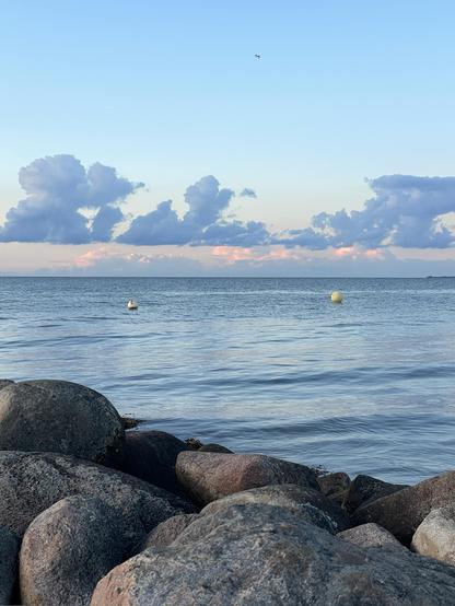 Eine ruhige Küstenszene mit ruhigem Wasser, vereinzelten Wolken am blauen Himmel und einer felsigen Küste im Vordergrund. Auf dem Wasser treiben zwei Bojen.