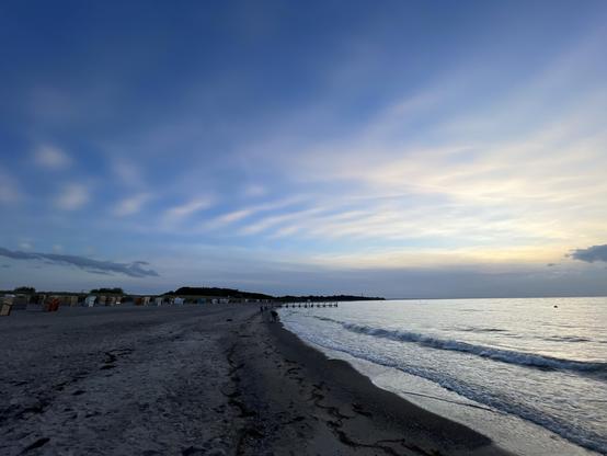Eine ruhige Strandszene bei Sonnenuntergang mit sandigem Ufer, sanften Wellen und einem farbenfrohen Himmel mit Wolkenstreifen. Im Hintergrund reihen sich Strandhütten aneinander, und ein Pier ragt ins Wasser, an dem ein paar Menschen entlanglaufen.