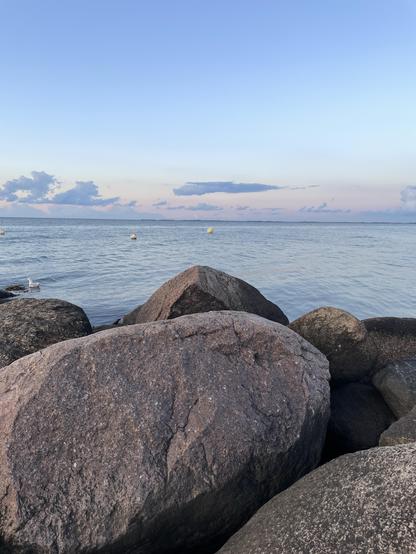 Eine Küstenszene mit großen Felsen im Vordergrund und ruhigem Wasser, das sich bis zum Horizont erstreckt. Weiche Wolken vor einem pastellfarbenen Himmel schaffen eine ruhige Atmosphäre, und auf dem Wasser sind in der Ferne einige Bojen zu sehen.
