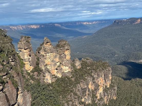 Echo Point, Australia Rock formation known as the Three Sisters, featuring three distinct sandstone peaks surrounded by lush green vegetation. The background includes a vast expanse of forested valleys and distant mountain ranges under a partly cloudy blue sky.
