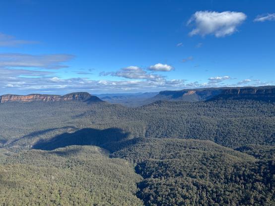 Jamison Valley, Australia Vast landscape of rolling green forested hills under a clear blue sky. A line of rugged cliffs is visible in the distance, with scattered white clouds above. Shadows from the clouds create darker patches on the hills below.